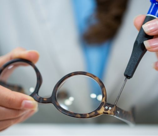 Nahaufnahme einer Person in einem Laborkittel, die mit einem Schraubenzieher eine braune Schildpattbrille repariert.