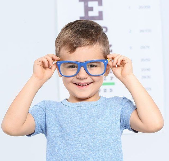 Ein kleines Kind in einem hellblauen Hemd hält sich blaue Ohrenschützer an die Ohren und steht vor einer Sehtafel in einer hellen Umgebung.